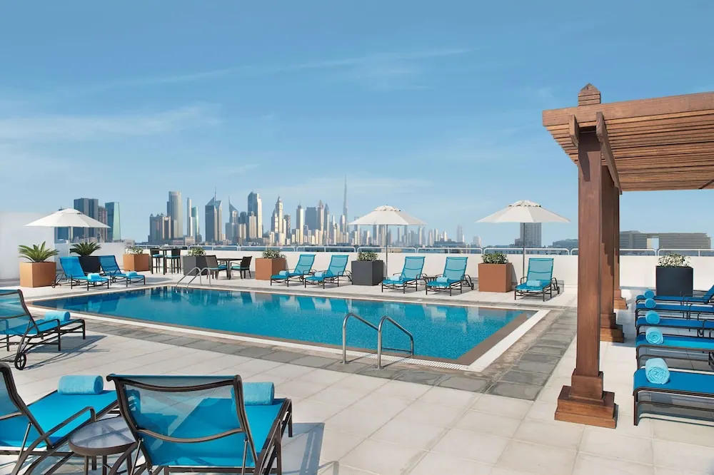 A rooftop swimming pool with blue loungers and white umbrellas, with the Dubai city skyline and the Burj Khalifa in the background.