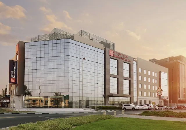 The exterior of a Hilton Garden Inn building with a glass facade, rooftop railings, a road in the foreground and parked cars at the side.