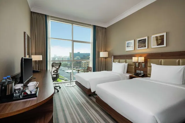 A twin bedroom with two white-bedded beds, a desk with a television and kettle tray, and floor-to-ceiling windows looking out over a city street and buildings.