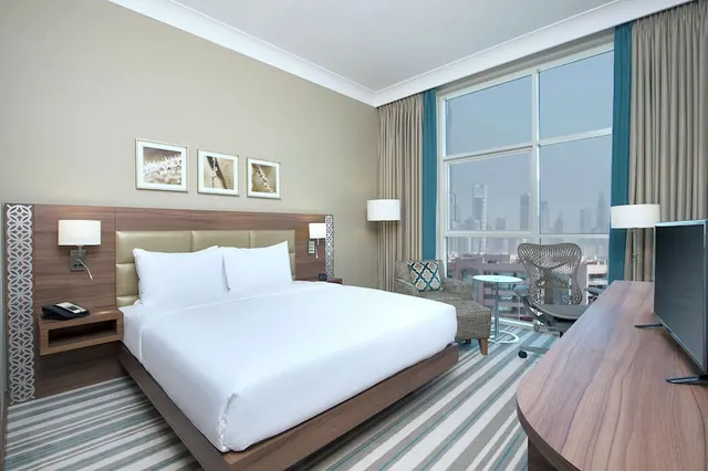 A hotel bedroom with a king bed, an armchair by a large window overlooking a hazy city skyline, and a wooden desk with a flat-screen television.