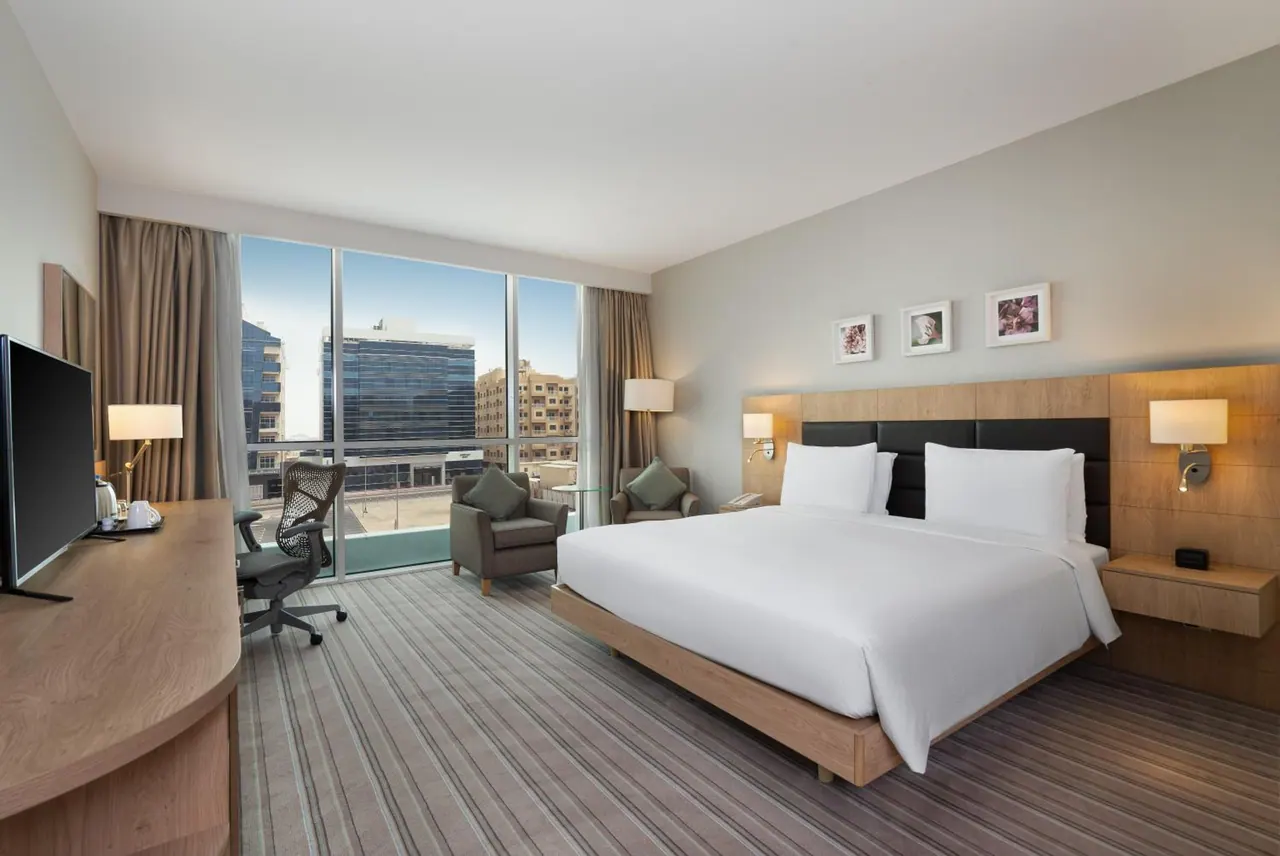 A king bed faces floor-to-ceiling windows with a city view, beside a work desk, armchairs and a wall-mounted television.
