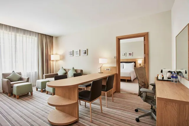 A hotel suite living area with a sofa, armchairs, a dining table with chairs, a desk with a kettle, and a view through an open doorway to a bedroom with a double bed.