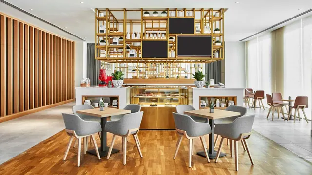A café dining area with grey upholstered chairs at wooden tables, a gold shelving display above a pastry counter, and tall windows on the right.