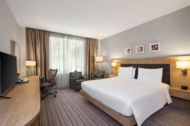 A hotel bedroom with a king bed with white linens, a wall-mounted television and desk with an office chair, and two armchairs beside a window with sheer curtains.