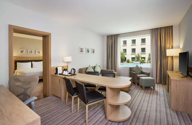 A hotel suite living area with a dining table, sofa and armchair, a wall-mounted television, and a bedroom visible through the doorway on the left, with a window overlooking a pool.
