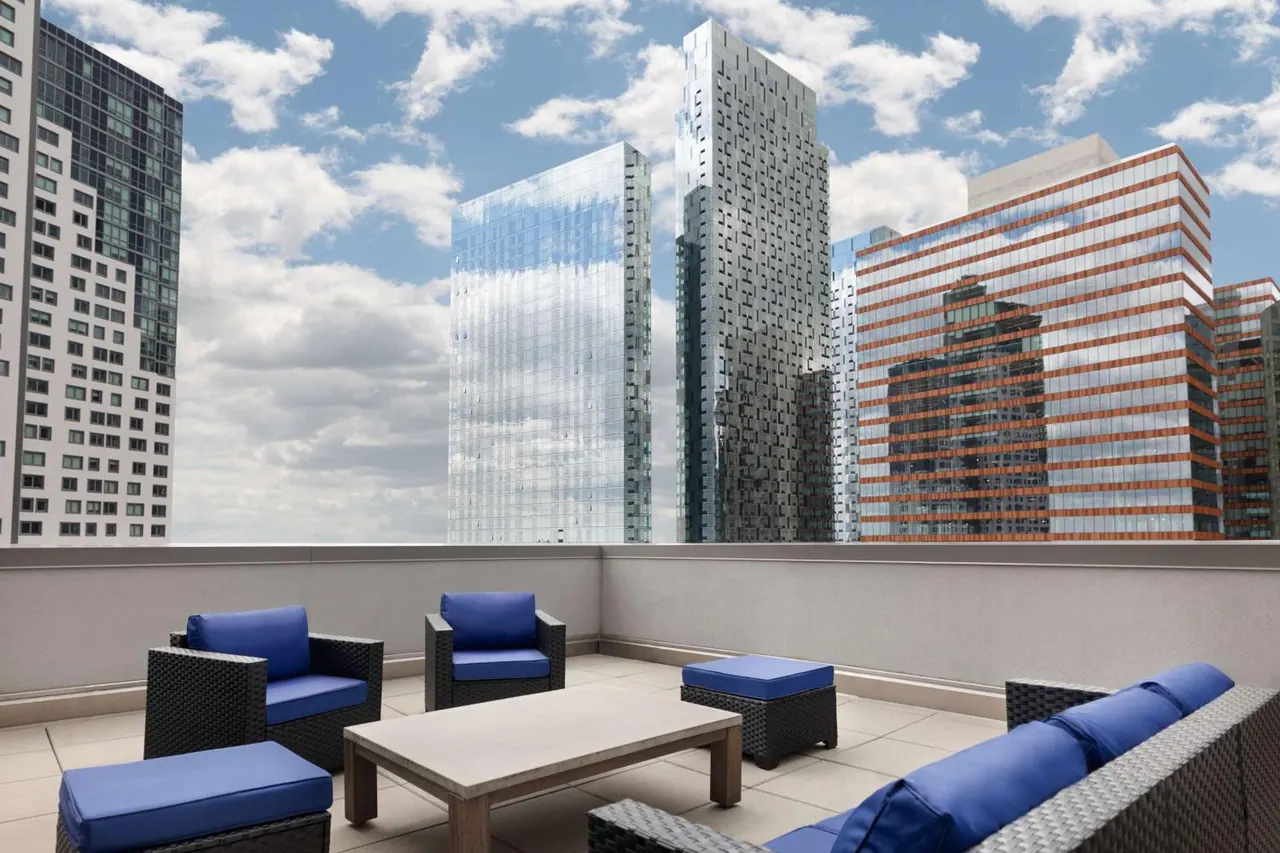 A rooftop terrace with wicker lounge chairs, blue cushions, a concrete coffee table and a view of high-rise buildings under a partly cloudy sky.