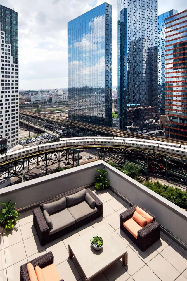 A furnished rooftop terrace with a wicker sofa, armchairs and a low table overlooks high-rise towers and an elevated railway.
