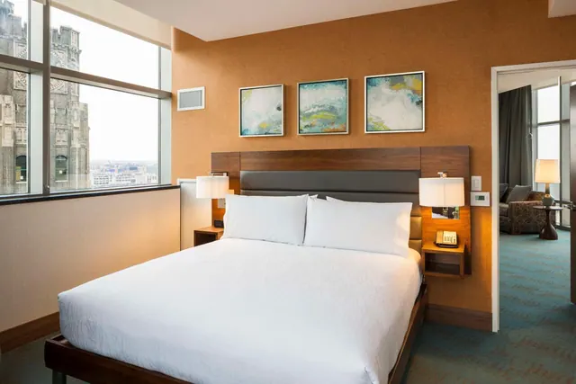 A king bed with white linens sits in a bedroom beside a large window overlooking a city with a gothic stone tower, and an open doorway shows a seating area in the next room.