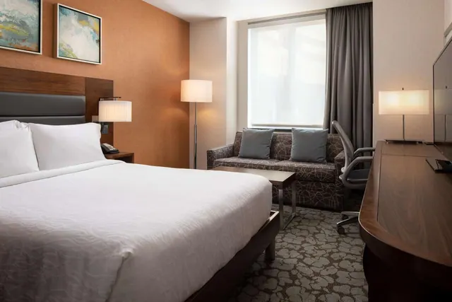 A king bed with white linens faces a grey patterned sofa with blue cushions, a wooden desk with a television, and a window in a hotel bedroom.