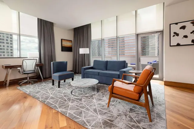 A living area with a blue sofa, blue and orange armchairs, a marble coffee table, and a desk by floor-to-ceiling windows overlooking city buildings and a balcony door.