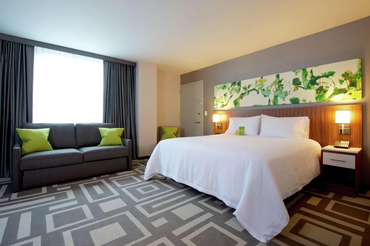 A hotel bedroom with a king bed, a grey sofa with green cushions beside a bright window, and a long green-and-white artwork above the headboard.