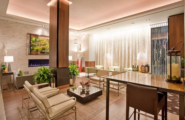 A lobby lounge with cream armchairs, a low dark coffee table, a fireplace set into a stone wall, potted plants, and a bar with bottles at the back.