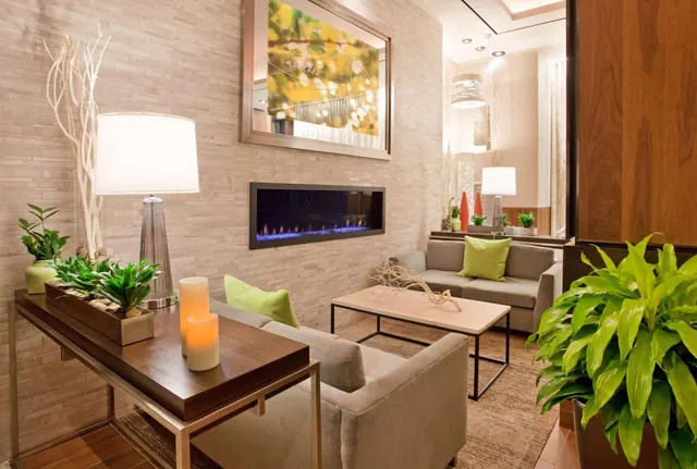 A lobby seating area with grey sofas, a low wooden-top coffee table, a wall-mounted fireplace, lit candles and potted plants.
