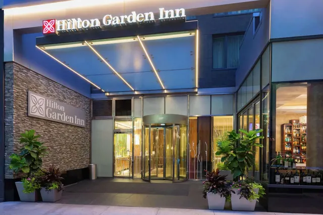 Entrance of a Hilton Garden Inn at dusk, with an illuminated canopy, a revolving door and potted plants on the pavement.
