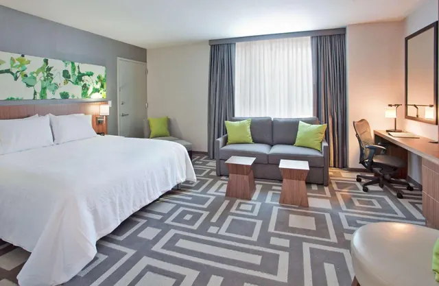 A hotel bedroom with a king bed, a grey sofa with green cushions, and a desk with an office chair beneath a wall mirror.
