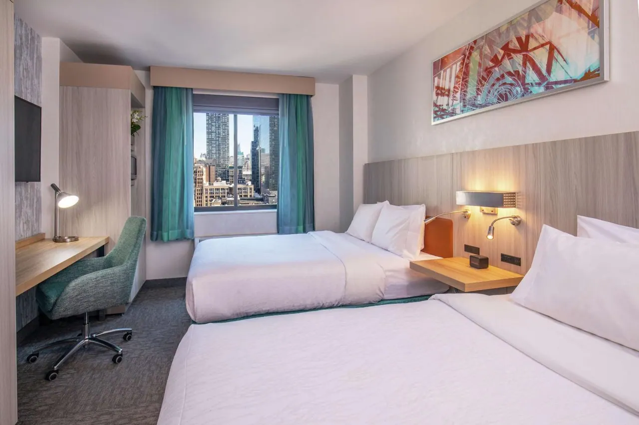 A hotel bedroom with two double beds, a desk and chair, a wall-mounted television, and a window looking out over city skyscrapers.