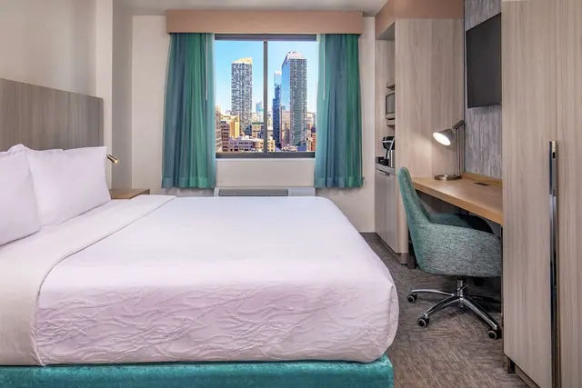 A bedroom with a king bed, a wall-mounted television, a desk with a lamp and chair, and a window overlooking city skyscrapers.