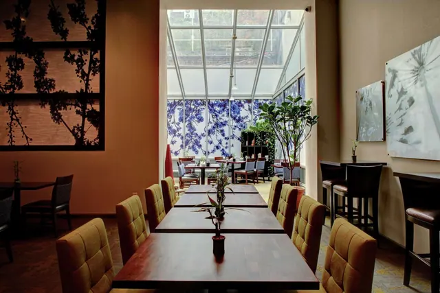 A dining room with rows of wooden tables and upholstered chairs, a glass-roofed atrium behind, and bar stools along the right wall.