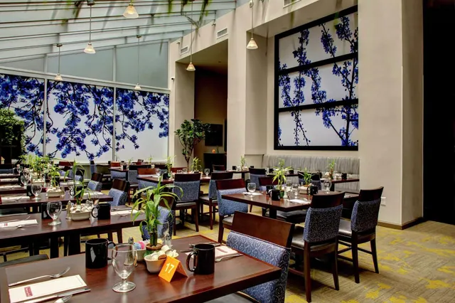 A restaurant dining room with set tables, blue patterned chairs, pendant lights, and large blue blossom artwork on the windows and wall.