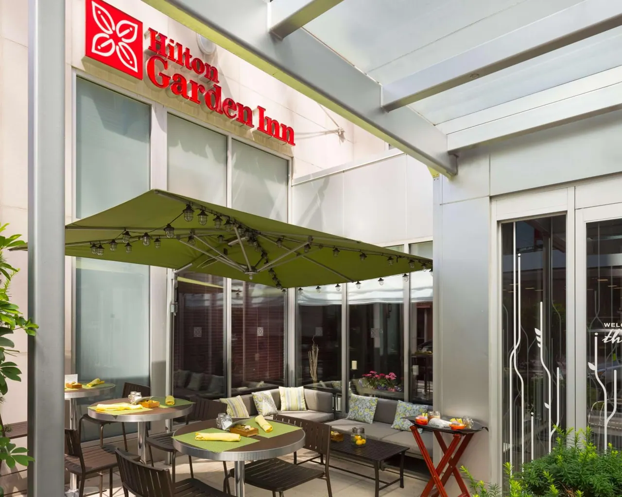 An outdoor patio with tables set under a large green umbrella, a sofa seating area, and a Hilton Garden Inn sign on the building behind.