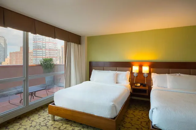 A hotel bedroom with two double beds beside floor-to-ceiling windows that open onto a balcony with a city skyline beyond.