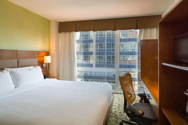 A made king bed with white linens sits beside a large window looking out on tall city buildings, with a wooden desk chair and wall-mounted television on the right.