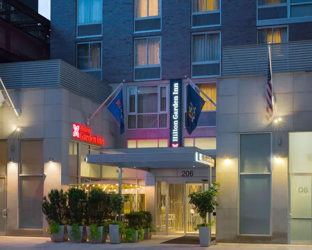 Exterior of a Hilton Garden Inn building at dusk, with a covered entrance, potted plants, and flags on the facade.