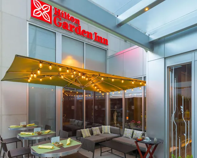 An outdoor terrace with a large canopy strung with lights, set tables with napkins, and a sofa seating area below the Hilton Garden Inn sign.
