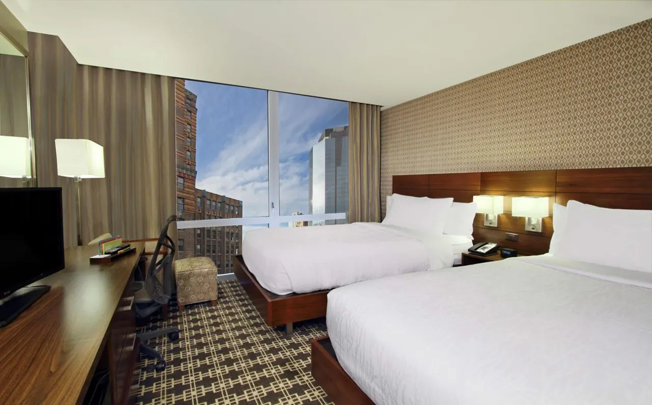 A hotel room with two made beds, a desk with a flat-screen TV, and a large window showing city buildings under a blue sky.