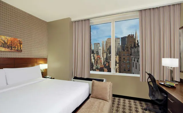 A bedroom with a king bed, a bench at the foot of the bed, a desk and chair, and a window looking out over city buildings including the Chrysler Building.