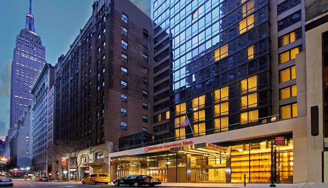 Exterior of a Hilton Garden Inn at dusk with lit windows, a yellow taxi and a black car on the street, and the Empire State Building visible in the background.