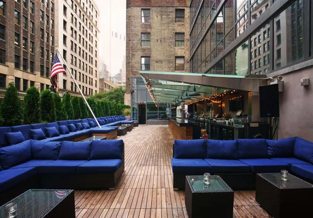 An outdoor terrace with blue sectional sofas, low glass tables, a wooden deck, and a bar with string lights under a glass canopy, surrounded by tall buildings.