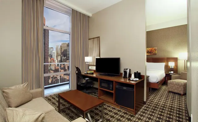 A hotel suite living area with a sofa, a desk holding a flat-screen TV and coffee machine, a tall window onto city high-rises, and a bed visible through a doorway.