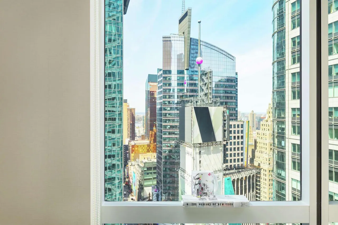 A window view over tall glass towers and a large billboard screen in a dense city district, with a small book on the windowsill.
