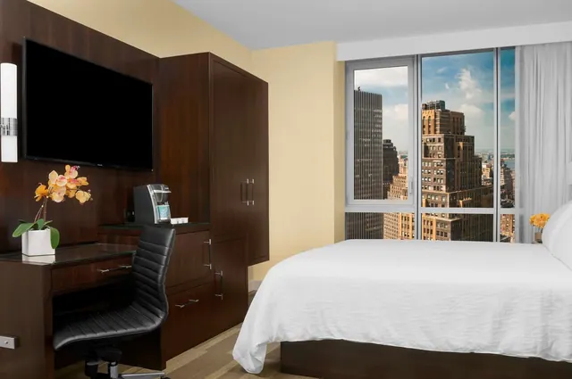 A bedroom with a king bed, wall-mounted television, desk with chair, and floor-to-ceiling windows looking out over city high-rises.