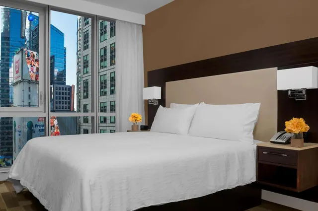 A king bed with white linens stands in a room with floor-to-ceiling windows that look out onto tall city buildings and billboards.
