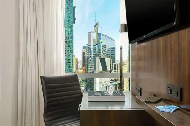 A work desk with a leather chair, a book, a glass object and a phone sits in front of a window showing city skyscrapers, beside a wall-mounted television.