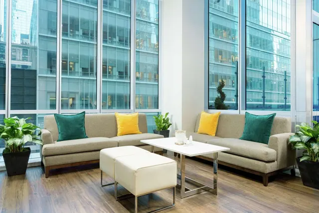 A seating area with two grey sofas, white ottomans and a white coffee table, with floor-to-ceiling windows overlooking glass office towers.