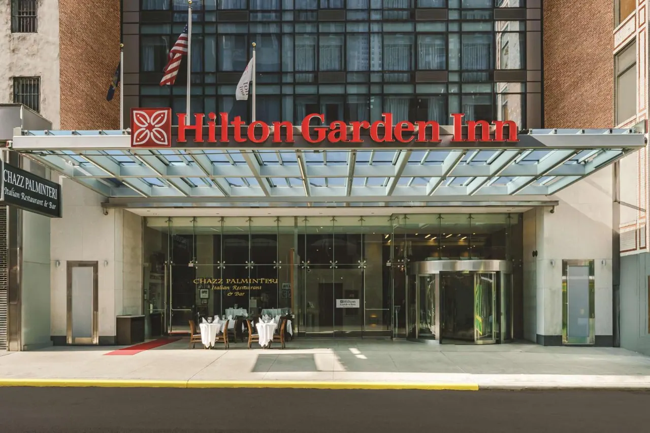 Front entrance of the Hilton Garden Inn under a glass canopy, with a revolving door and the Chazz Palmintieri restaurant with set tables visible behind the glass.