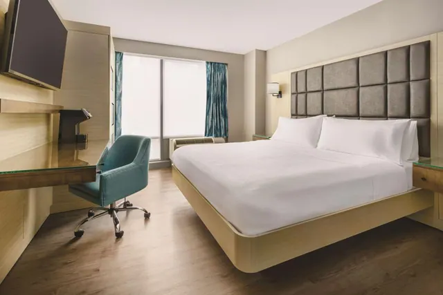 A bedroom with a large king bed with white linens, a padded grey headboard, a teal desk chair beside a wooden desk, and a wall-mounted television.
