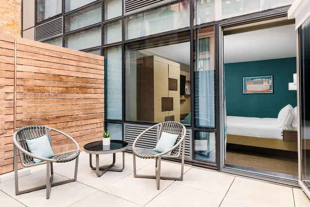 A private terrace with two grey woven chairs, a small round side table with a potted plant, and a wooden slatted wall, with a glass sliding door opening into a bedroom with a teal accent wall and a bed.