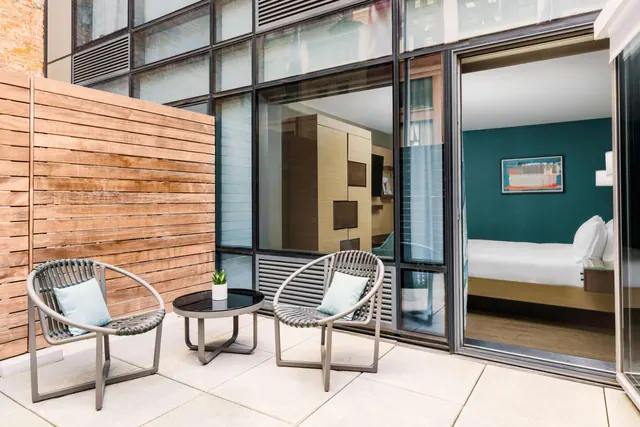 A private terrace with two grey woven chairs, a small round side table with a plant, and a wooden slatted privacy wall, with a bedroom visible through the glass sliding door.