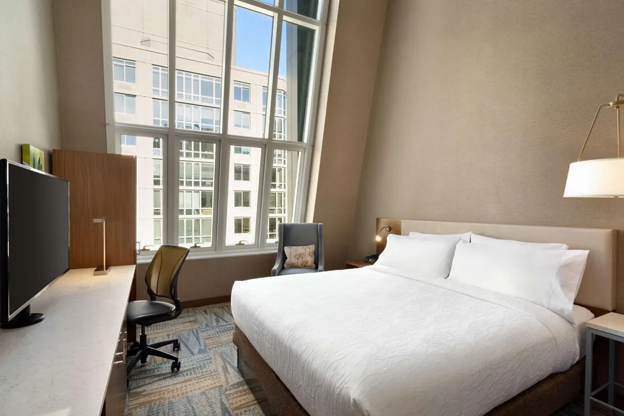 A hotel bedroom with a king bed, a desk with a wall-mounted television, and a tall window looking out onto a neighbouring building under a blue sky.