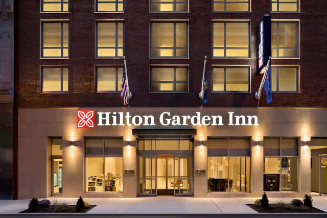 Exterior of a brick Hilton Garden Inn building at dusk, with lit windows, an illuminated entrance and flags on the facade.