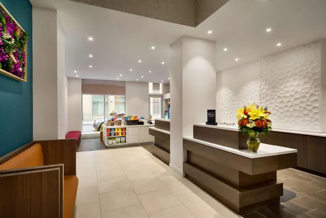 A hotel lobby with a wooden reception desk, a vase of flowers, an orange bench seat, and a street with a parked car visible through the windows at the back.