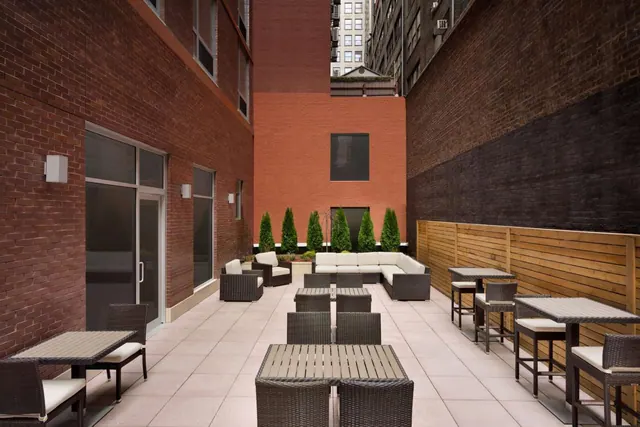 An outdoor terrace with wicker dining tables and chairs and a sectional sofa group, enclosed by brick walls and a wooden fence with shrubs along the back wall.
