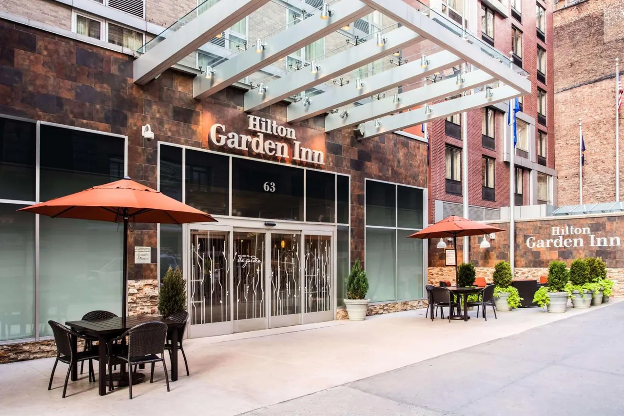 The Hilton Garden Inn entrance has a glass canopy, outdoor tables under orange umbrellas, and potted plants along the sidewalk.
