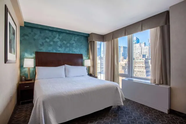 A bedroom with a king bed, a teal patterned accent wall, and a window looking out over city skyscrapers.