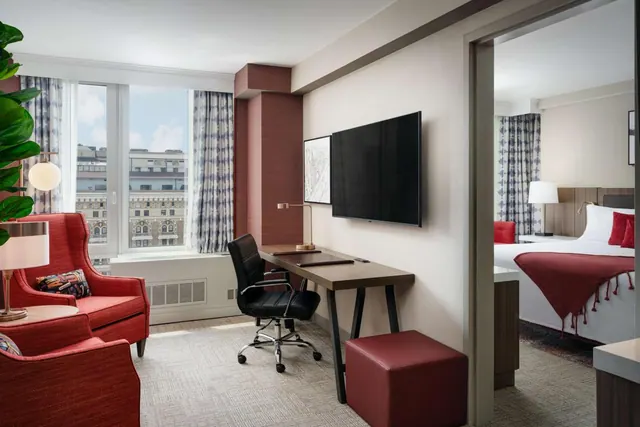 A living area with two red armchairs, a desk with a wall-mounted television, and a view through the doorway to a bed with a red throw, with city buildings visible through the window.
