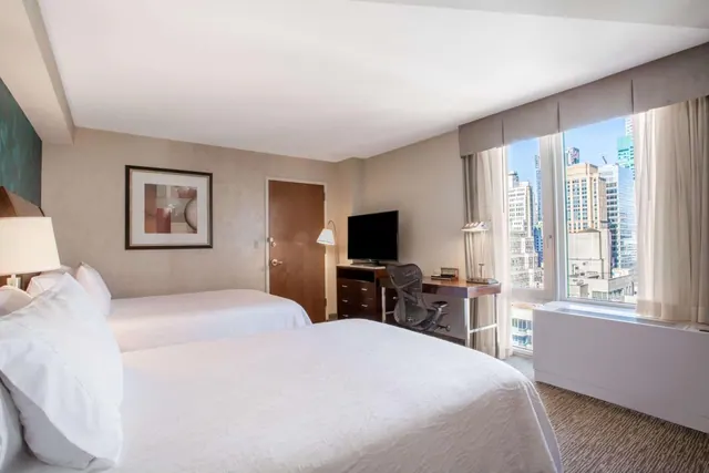 A hotel bedroom with two beds, a desk with a chair, a wall-mounted television, and a large window looking out over city buildings.